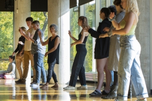 A group of nine people stand in a line practicing tai chi in a well-lit studio