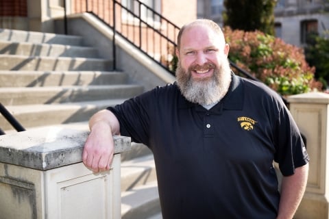Chuck Xander, a light-skinned man with a full, graying beard, smiles for a photo wearing a black University of Iowa polo shirt.
