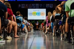 A shot down an aisle of students watching a presentation. At the front, a man stands in front of a podium and behind him a projection of a presentation about free speech, including a four-way Venn diagram, is shown.