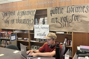 A student in a ball cap sits at a laptop with the words "The George and Mary Foster Anthropology Library" on official lettering on the wall behind him, and, on banners below those letters, “You fund displacement why not education?” and “public libraries are not for sale." 