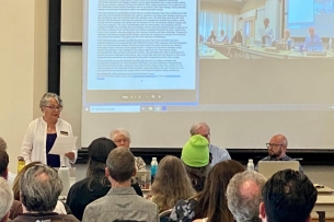 Christina Cuevas, chair of the Name Exploration Subcommittee, stands and speaks while her fellow board members sit next to her in front of a crowd of people.