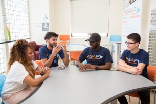 Four students sit around a table talking. Three of them are wearing navy blue shirts that read "ASAP" in blue and orange letters.
