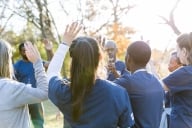 A group of students in dark blue shirts raise their hands outside in the fall as part of some kind of group event.