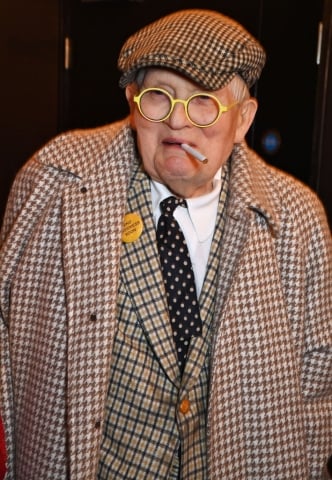 A photo of a gentleman in a checkered coat, plaid jacket, tie, and cap. He has large round eyeglasses and a cigarette in his mouth.