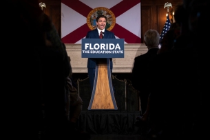 Ron DeSantis stands at a lectern that says "Florida The Education State," with the state flag behind him.