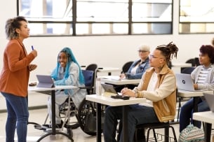 A professor stands in front of a group of students who are sitting at desks. 