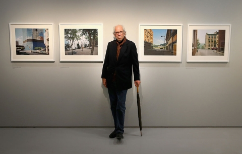 A man in a black blazer uses an umbrella as a cane and stands in front of four framed photographs.