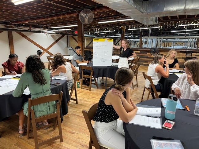 Students sit at round tables, listening to a peer present from a posterboard