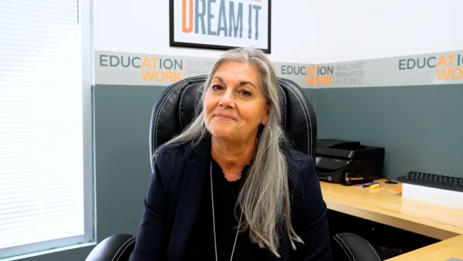 Jane Swift, a light-skinned woman with long gray hair wearing a black shirt, sits at her desk at the Education at Work headquarters in Arizona.