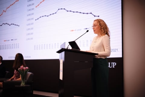 A woman speaks at a podium in front of a graph on a projector screen.