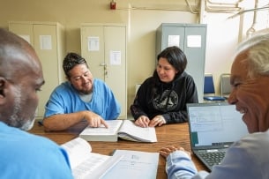 A student in a blue jumpsuit points to a book, smiling, with an instructor sitting beside him.