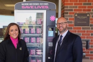 Two individuals stand next to a pink-and-black vending machine containing Narcan.