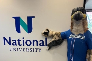 A person wearing a falcon mascot costume poses next to a sign reading "National University."