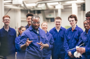 A group of engineering students in blue coveralls listens to a professor.