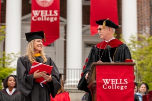 Wells College president Jonathan Gibralter, a light-skinned man with gray hair and a mustache, wearing academic regalia and sunglasses, on graduation day in front of red Wells College signage.