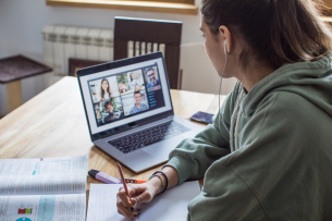 A woman in a sweatshirt leans over a table working with papers and looking at a laptop with other people on the screen