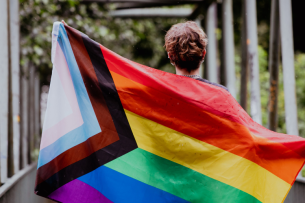 A young person carries a Progress Pride flag unfurled behind their back.