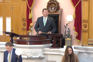 A man in a suit stands at a raised podium above another man and a woman, backed by gold and red curtains.