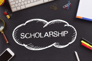 A graphic with the word "scholarship" in a cloud-shaped bubble, against the background of a desk, with a keyboard and school supplies visible in the background.