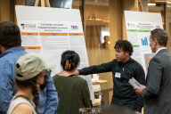 Two students in a large room share their science research poster with a professor as others walk by.