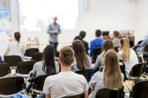 Students sit in a lecture hall facing the instructor at the front of the room